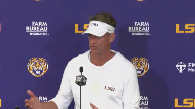 LSU coach at a press conference podium, wearing a white hoodie and visor, speaking against a purple LSU backdrop with logos.