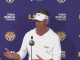 LSU coach at a press conference podium, wearing a white hoodie and visor, speaking against a purple LSU backdrop with logos.