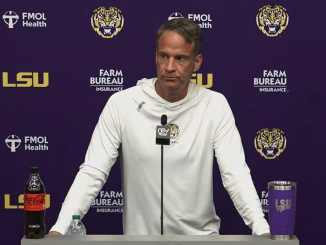 Male LSU coach at a press conference podium with microphone, wearing a white hoodie; purple backdrop with LSU and sponsor logos and drinks on the table (Coca-Cola bottle left, tumbler right).
