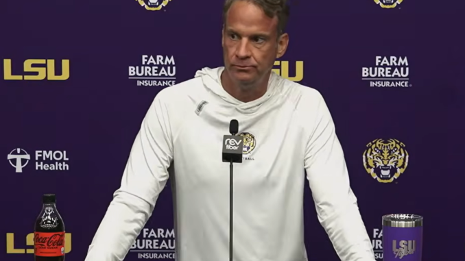 Male LSU coach at a press conference podium with microphone, wearing a white hoodie; purple backdrop with LSU and sponsor logos and drinks on the table (Coca-Cola bottle left, tumbler right).