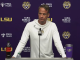 Male LSU coach at a press conference podium with microphone, wearing a white hoodie; purple backdrop with LSU and sponsor logos and drinks on the table (Coca-Cola bottle left, tumbler right).