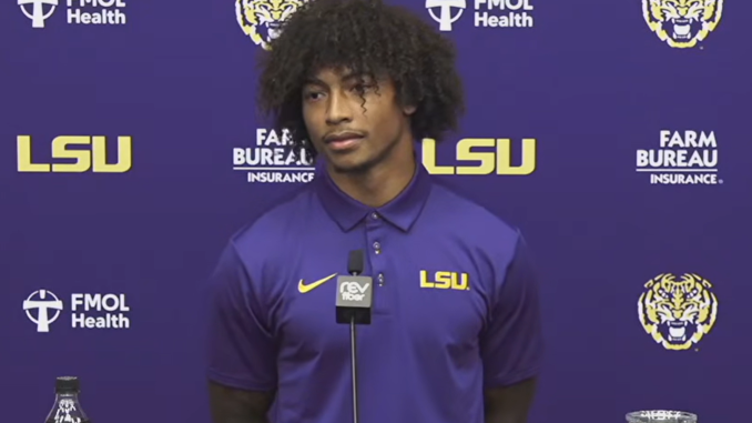 Young man in a purple LSU polo stands at a podium with a microphone during a press conference backdrop.