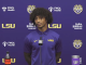 Young man in a purple LSU polo stands at a podium with a microphone during a press conference backdrop.