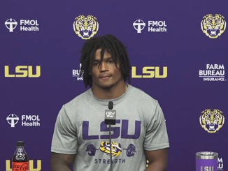 LSU athlete at a press conference in a purple backdrop with LSU logos, speaking into a microphone, wearing a gray LSU shirt.