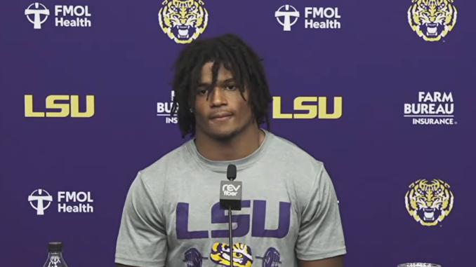 LSU athlete at a press conference in a purple backdrop with LSU logos, speaking into a microphone, wearing a gray LSU shirt.