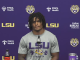 LSU athlete at a press conference in a purple backdrop with LSU logos, speaking into a microphone, wearing a gray LSU shirt.