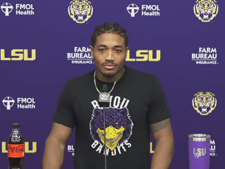 Athlete at a LSU press conference wearing a black shirt with a tiger graphic, microphone at center, purple backdrop with LSU and sponsor logos behind him.