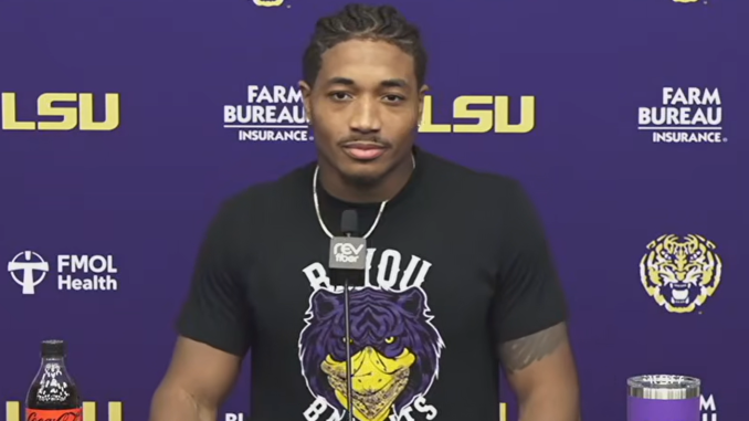 Athlete at a LSU press conference wearing a black shirt with a tiger graphic, microphone at center, purple backdrop with LSU and sponsor logos behind him.