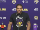 Athlete at a LSU press conference wearing a black shirt with a tiger graphic, microphone at center, purple backdrop with LSU and sponsor logos behind him.