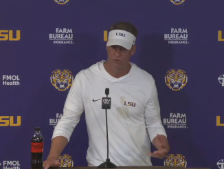 Man in white LSU hoodie and visor speaks at a podium in a LSU backdrop with sponsor logos; Coca-Cola bottle on the table nearby