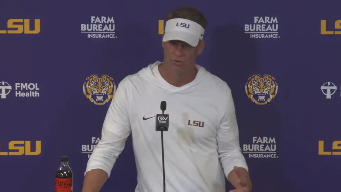 Man in white LSU hoodie and visor speaks at a podium in a LSU backdrop with sponsor logos; Coca-Cola bottle on the table nearby
