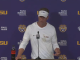 Man in white LSU hoodie and visor speaks at a podium in a LSU backdrop with sponsor logos; Coca-Cola bottle on the table nearby