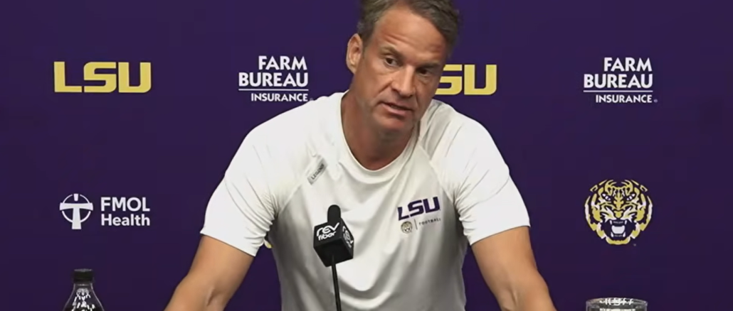 Press conference scene with LSU coach at a podium, purple backdrop featuring LSU, FMOL Health, and Farm Bureau logos; coach in white LSU shirt speaks into a microphone.