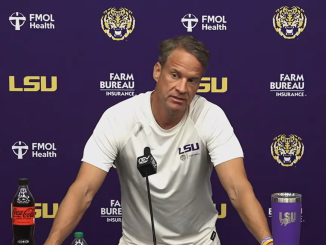Press conference scene with LSU coach at a podium, purple backdrop featuring LSU, FMOL Health, and Farm Bureau logos; coach in white LSU shirt speaks into a microphone.