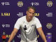 Press conference scene with LSU coach at a podium, purple backdrop featuring LSU, FMOL Health, and Farm Bureau logos; coach in white LSU shirt speaks into a microphone.