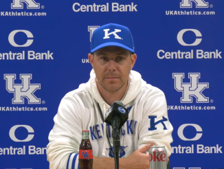 Kentucky coach at a press conference in front of a blue UK backdrop with Central Bank logos, wearing a blue cap and white hoodie, microphone in front.
