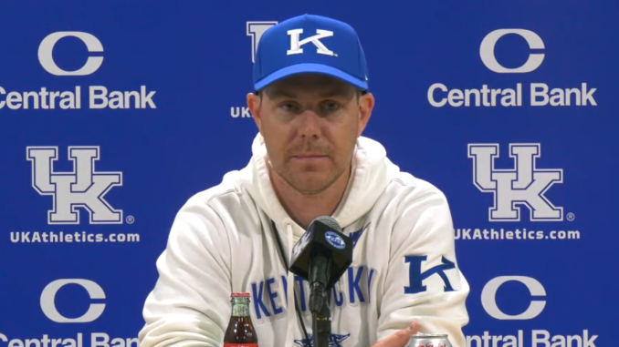 Kentucky coach at a press conference in front of a blue UK backdrop with Central Bank logos, wearing a blue cap and white hoodie, microphone in front.
