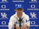 Kentucky coach at a press conference in front of a blue UK backdrop with Central Bank logos, wearing a blue cap and white hoodie, microphone in front.