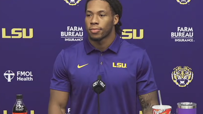 LSU athlete in a purple polo speaks at a press conference with Coca‑Cola bottle and Cane's cup on the table, LSU logos in the background.
