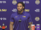 LSU athlete in a purple polo speaks at a press conference with Coca‑Cola bottle and Cane's cup on the table, LSU logos in the background.