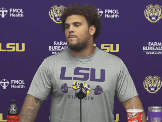 LSU athlete in a gray shirt at a press conference, standing before a purple backdrop with LSU logos and sponsors; Coca‑Cola bottle and a Cane’s cup on the table.