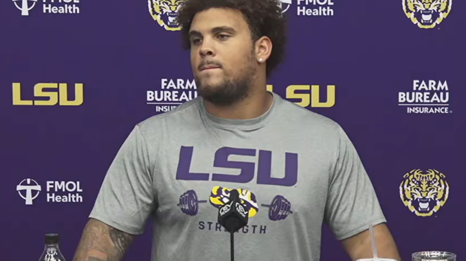 LSU athlete in a gray shirt at a press conference, standing before a purple backdrop with LSU logos and sponsors; Coca‑Cola bottle and a Cane’s cup on the table.