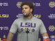 LSU athlete in a gray shirt at a press conference, standing before a purple backdrop with LSU logos and sponsors; Coca‑Cola bottle and a Cane’s cup on the table.
