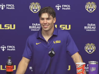 LSU athlete at a press conference, smiling in a purple LSU polo with sponsor logos in the background.