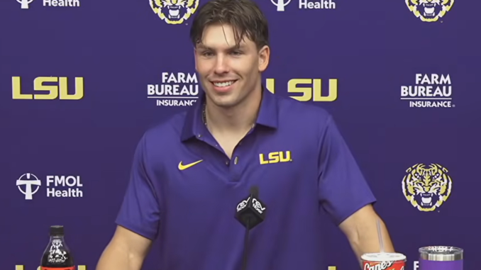 LSU athlete at a press conference, smiling in a purple LSU polo with sponsor logos in the background.