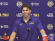 LSU athlete at a press conference, smiling in a purple LSU polo with sponsor logos in the background.