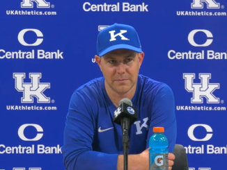 Male Kentucky coach wearing a blue cap with a K, speaking at a press conference with a blue UK backdrop featuring Central Bank logos and UK Athletics.