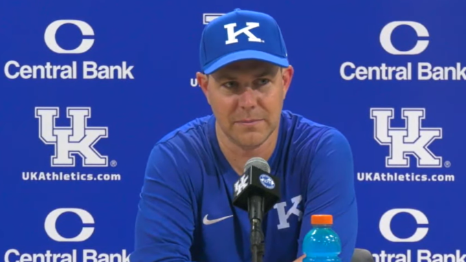 Male Kentucky coach wearing a blue cap with a K, speaking at a press conference with a blue UK backdrop featuring Central Bank logos and UK Athletics.