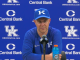 Male Kentucky coach wearing a blue cap with a K, speaking at a press conference with a blue UK backdrop featuring Central Bank logos and UK Athletics.