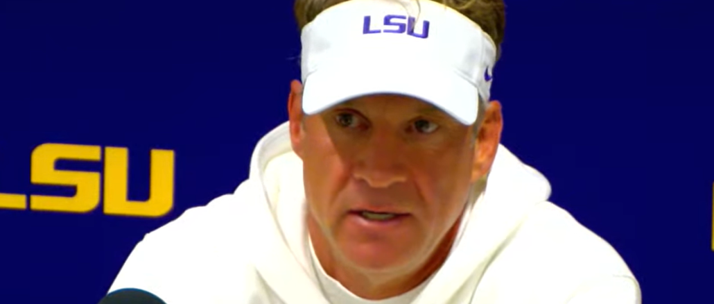 LSU coach in a white hoodie and visor speaks at a Lexington press conference with microphones in front of him.