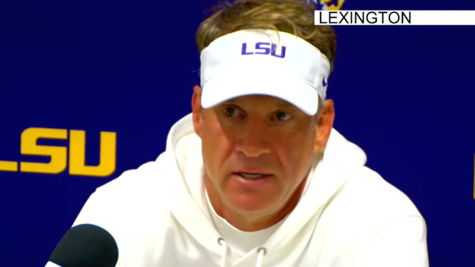 LSU coach in a white hoodie and visor speaks at a Lexington press conference with microphones in front of him.