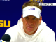 LSU coach in a white hoodie and visor speaks at a Lexington press conference with microphones in front of him.