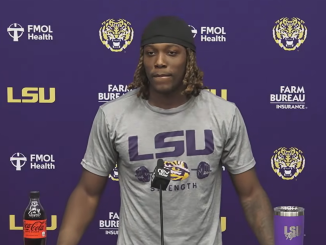 LSU athlete at a press conference podium wearing a gray LSU shirt, with a purple backdrop and tiger logos behind him