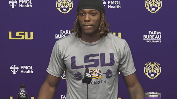 LSU athlete at a press conference podium wearing a gray LSU shirt, with a purple backdrop and tiger logos behind him