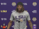 LSU athlete at a press conference podium wearing a gray LSU shirt, with a purple backdrop and tiger logos behind him