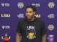 Young man at a LSU press conference against a purple backdrop with sponsor logos; a Coke bottle and LSU tumbler on the table.