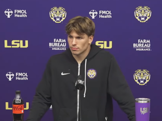 Young man in a black hoodie at a LSU press conference backdrop with team logos and sponsor names visible (Coca-Cola bottle on table).
