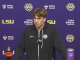 Young man in a black hoodie at a LSU press conference backdrop with team logos and sponsor names visible (Coca-Cola bottle on table).