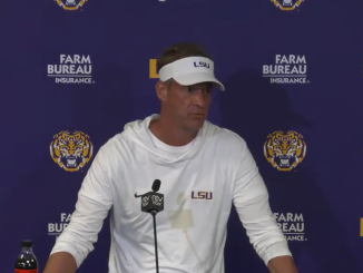 Man in a white LSU hoodie and visor speaks at a press conference in front of a Farm Bureau backdrop with tiger logos behind him
