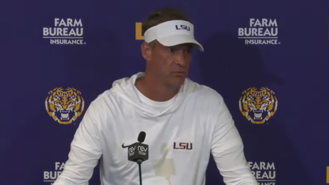 Man in a white LSU hoodie and visor speaks at a press conference in front of a Farm Bureau backdrop with tiger logos behind him