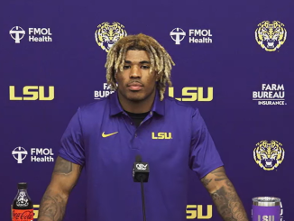 Athlete in a purple LSU polo speaks at a press conference, standing behind a mic with a purple backdrop and LSU logos far and wide.