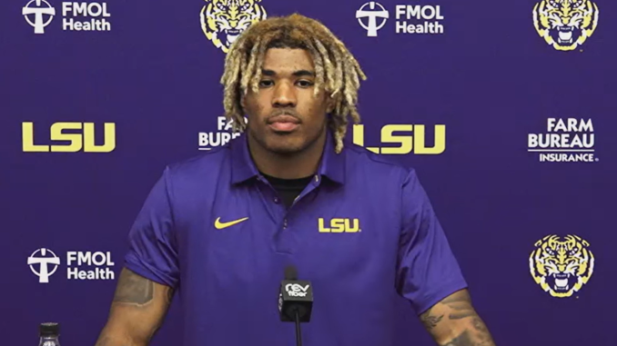 Athlete in a purple LSU polo speaks at a press conference, standing behind a mic with a purple backdrop and LSU logos far and wide.
