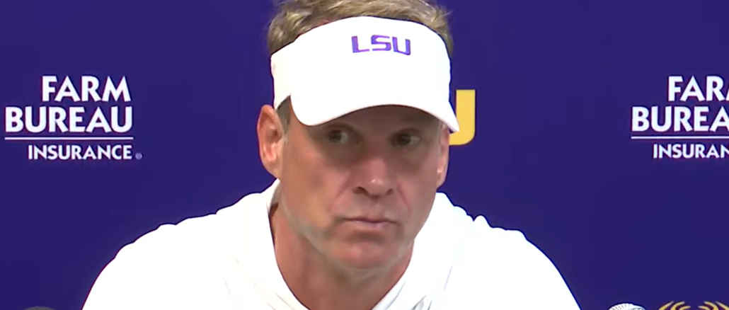 Man in a white LSU visor speaks at a press conference, microphones in front with a blue backdrop showing Farm Bureau Insurance logos and Auburn text.