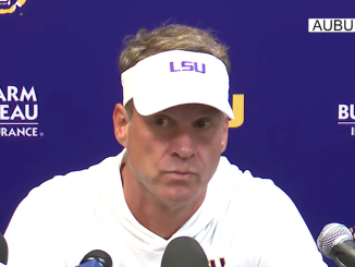 Man in a white LSU visor speaks at a press conference, microphones in front with a blue backdrop showing Farm Bureau Insurance logos and Auburn text.