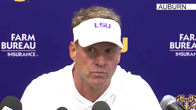 Man in a white LSU visor speaks at a press conference, microphones in front with a blue backdrop showing Farm Bureau Insurance logos and Auburn text.
