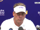 Man in a white LSU visor speaks at a press conference, microphones in front with a blue backdrop showing Farm Bureau Insurance logos and Auburn text.